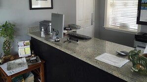 A reception desk with a computer, printer, and keyboard. There is a clock on the wall and a window with blinds. - Roswell Inn (Roswell)