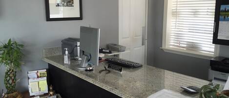 A reception desk with a computer, printer, and keyboard. There is a clock on the wall and a window with blinds.