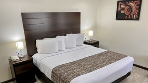 A neatly made bed with white linens and a patterned throw, flanked by two bedside tables with lamps, in a room with a dark wood headboard and a single artwork on the wall. - Roswell Inn (Roswell)