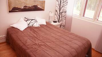 A bedroom with a large bed, a bedside table with a lamp, a wall-mounted painting, and a window with blinds.