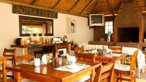 A rustic dining area with wooden tables and chairs, a brick fireplace, and a television mounted on the wall. - Boma Lodge (Durban North)