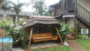 A wooden swing with a thatched roof in a garden area with a pool and potted plants. - Boma Lodge (Durban North)
