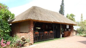 A thatched-roof building with a brick exterior, a wooden deck, and a small garden area with plants and a birdbath. - Boma Lodge (Durban North)