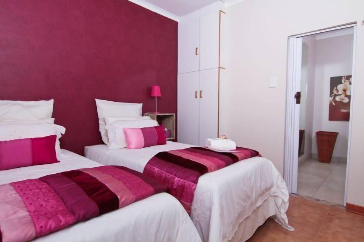 A hotel room with two beds, each with white bedding and pink accents. There is a bedside table with a lamp, and a wardrobe with a mirror.