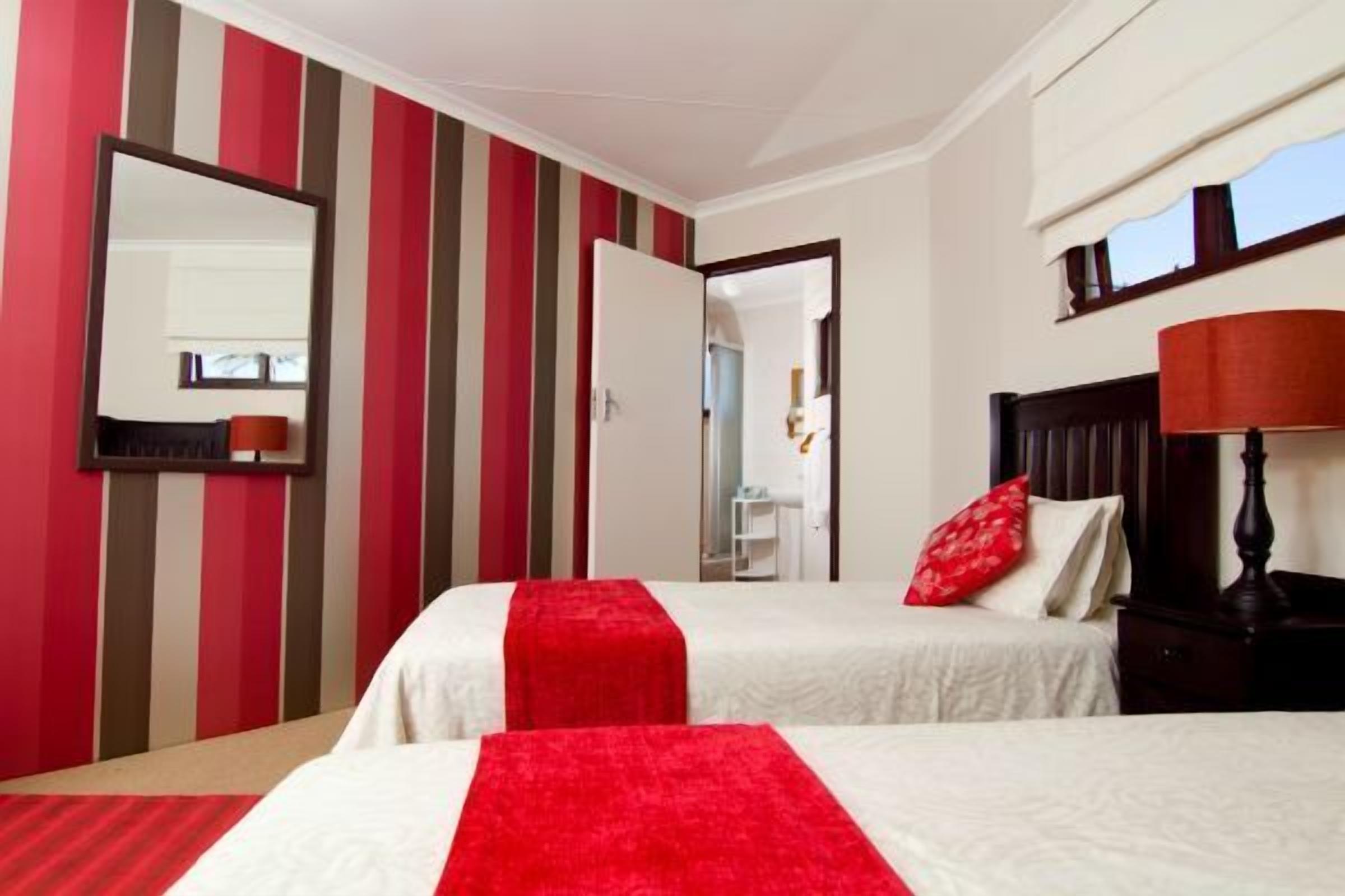 A hotel room with striped walls, two beds with red and white bedding, a wooden nightstand, a mirror, and a window with a view.