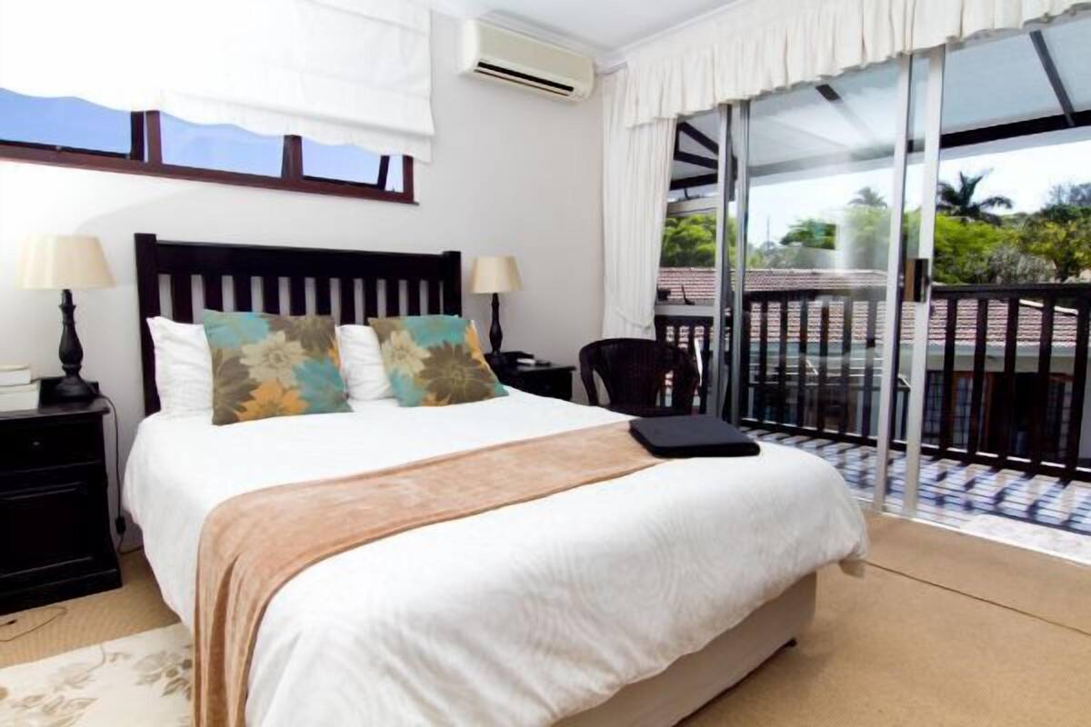 A hotel room with a large bed, two bedside tables with lamps, a chair, a balcony with a table and chairs, and a view of greenery.