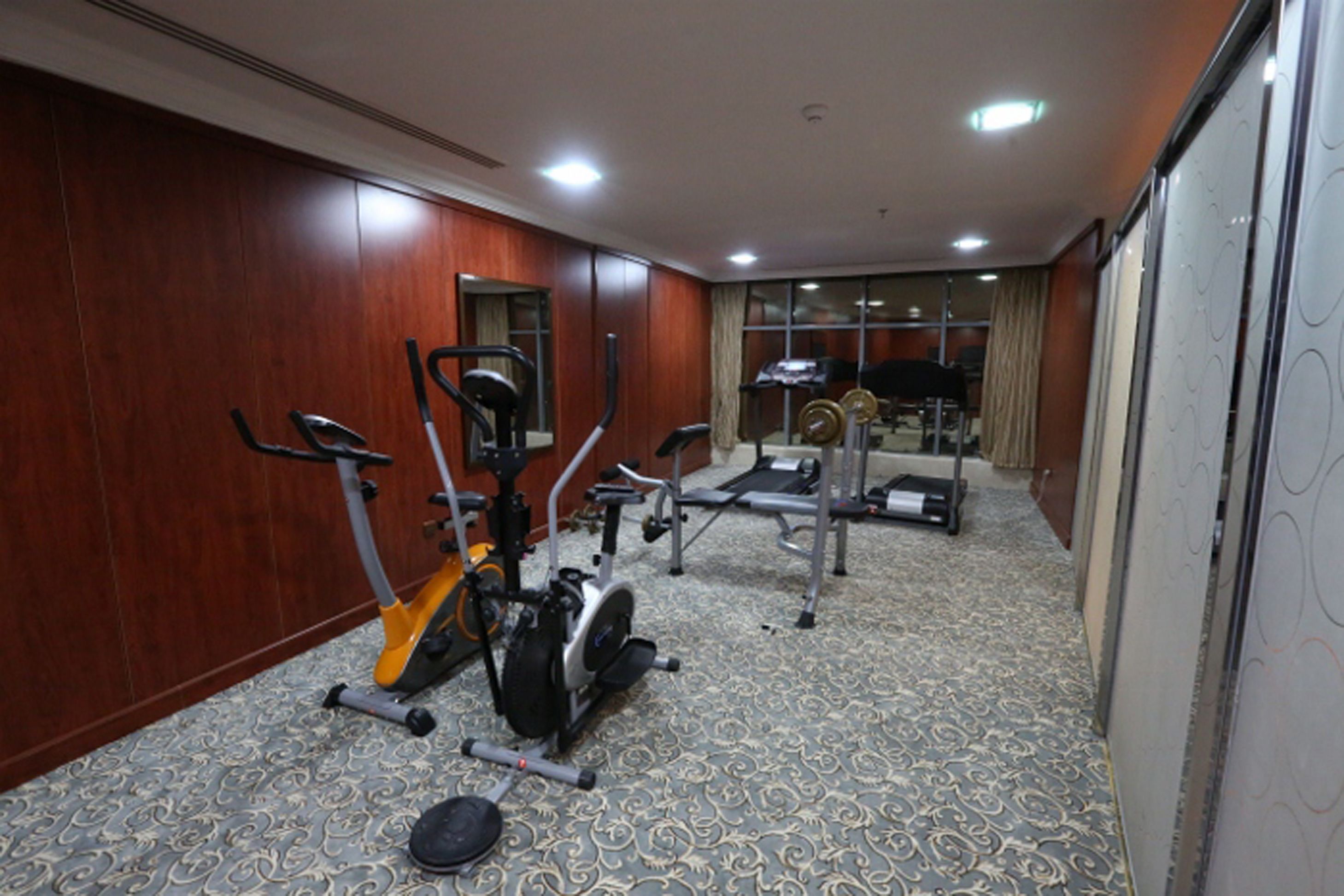 Habitación de hotel con una amplia zona de ejercicio que incluye equipo de cardio y pesas, un comedor con mesa y sillas, y un gran ventanal con cortinas.