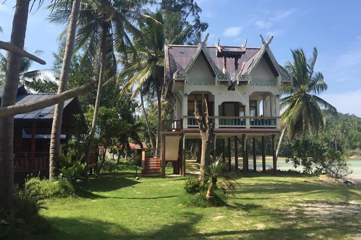 Uma casa de dois andares com varanda, rodeada por palmeiras e uma praia.