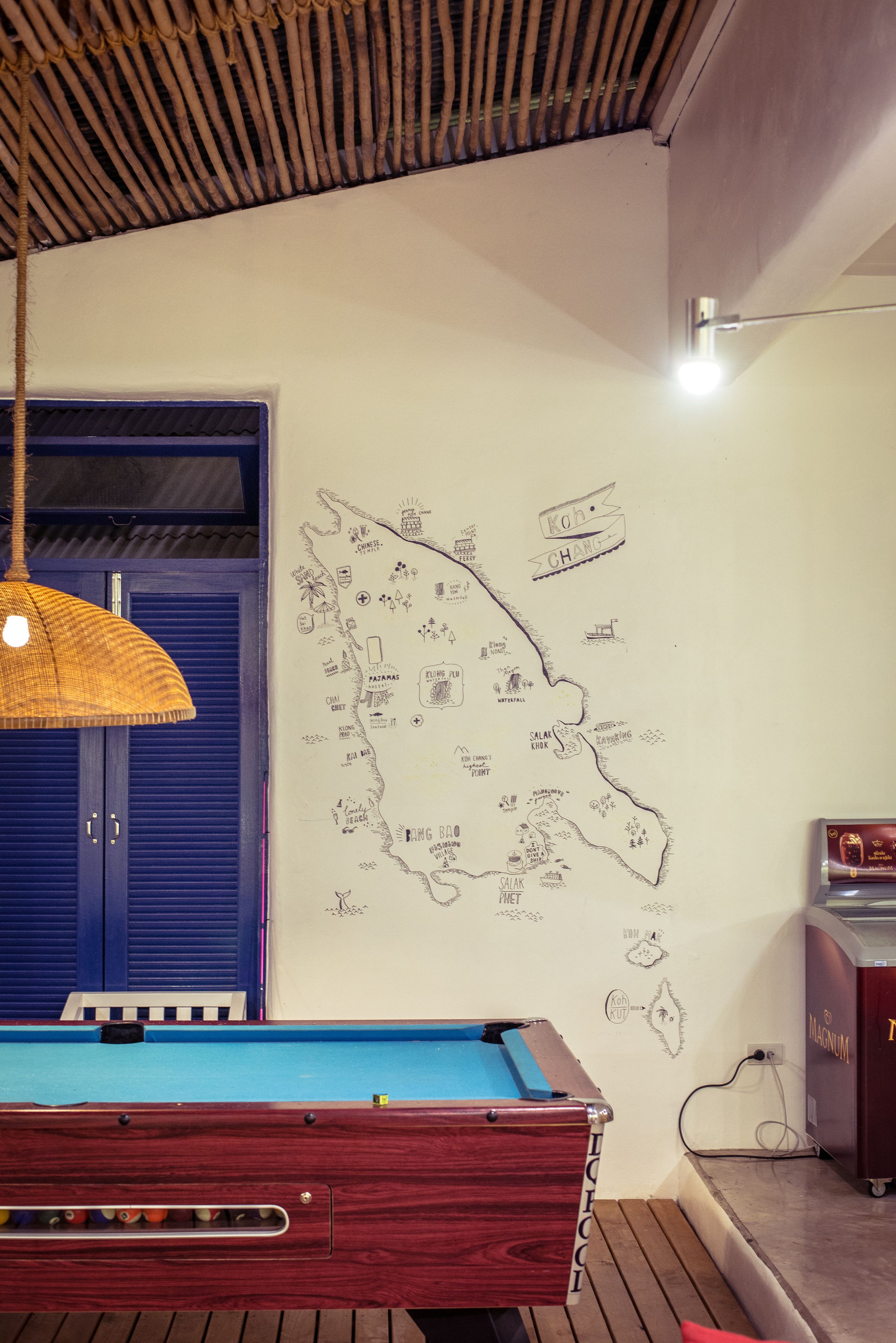 Una mesa de billar con acabado de madera y superficie de fieltro azul, una pared con un mapa y varios iconos, una puerta azul y una lámpara colgante.