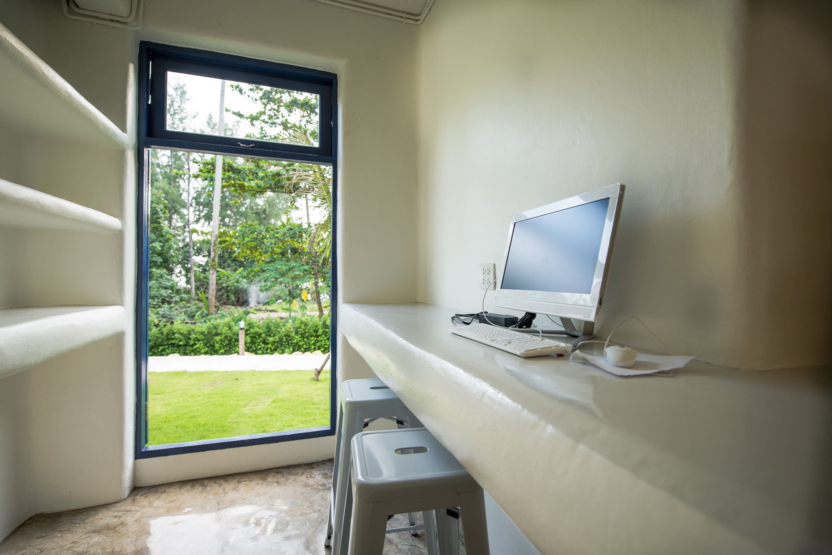 A room with a desk, a computer, and a window with a view of greenery.