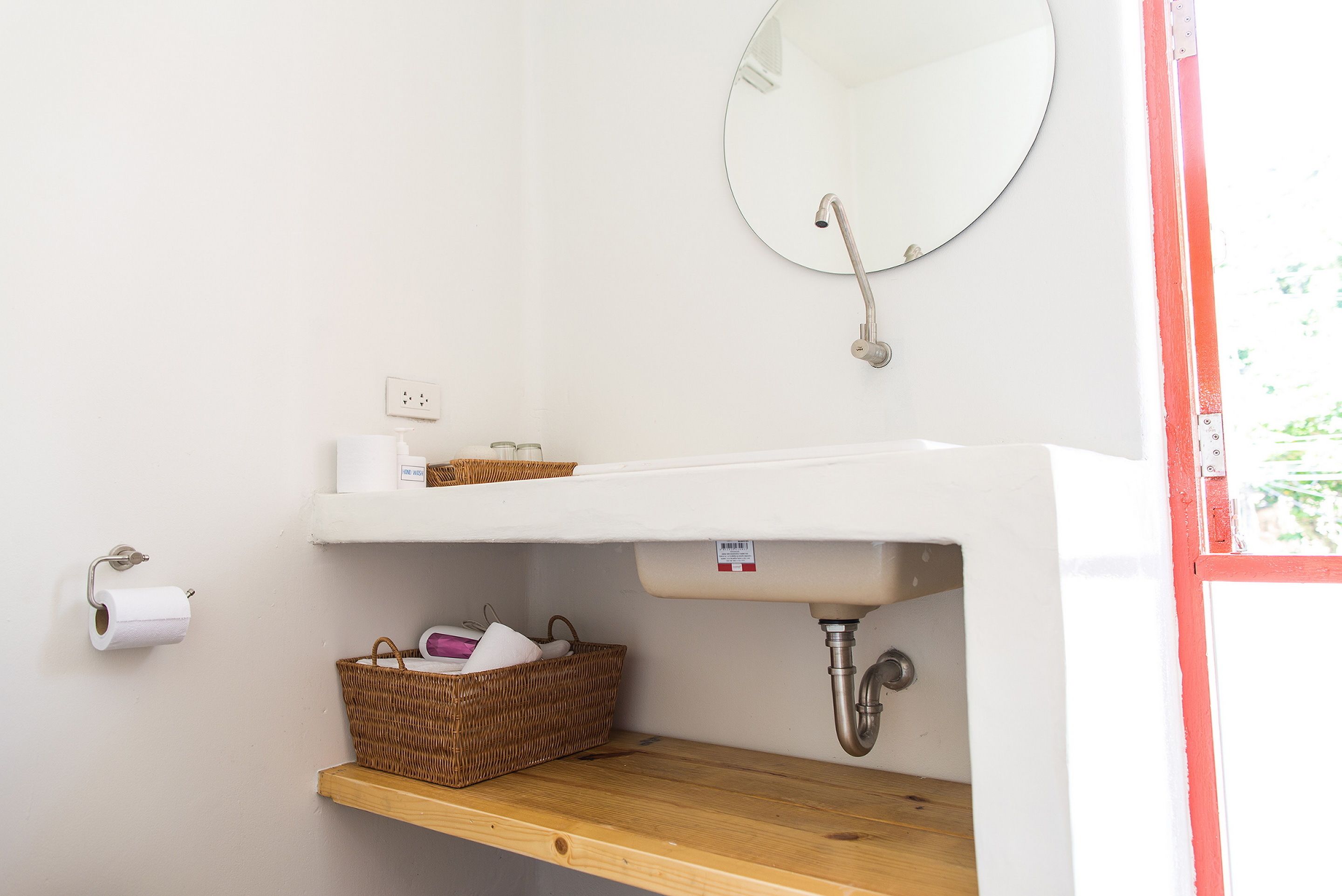 A bathroom with a white sink, a mirror, a wooden shelf, a wicker basket, and a toilet paper holder.