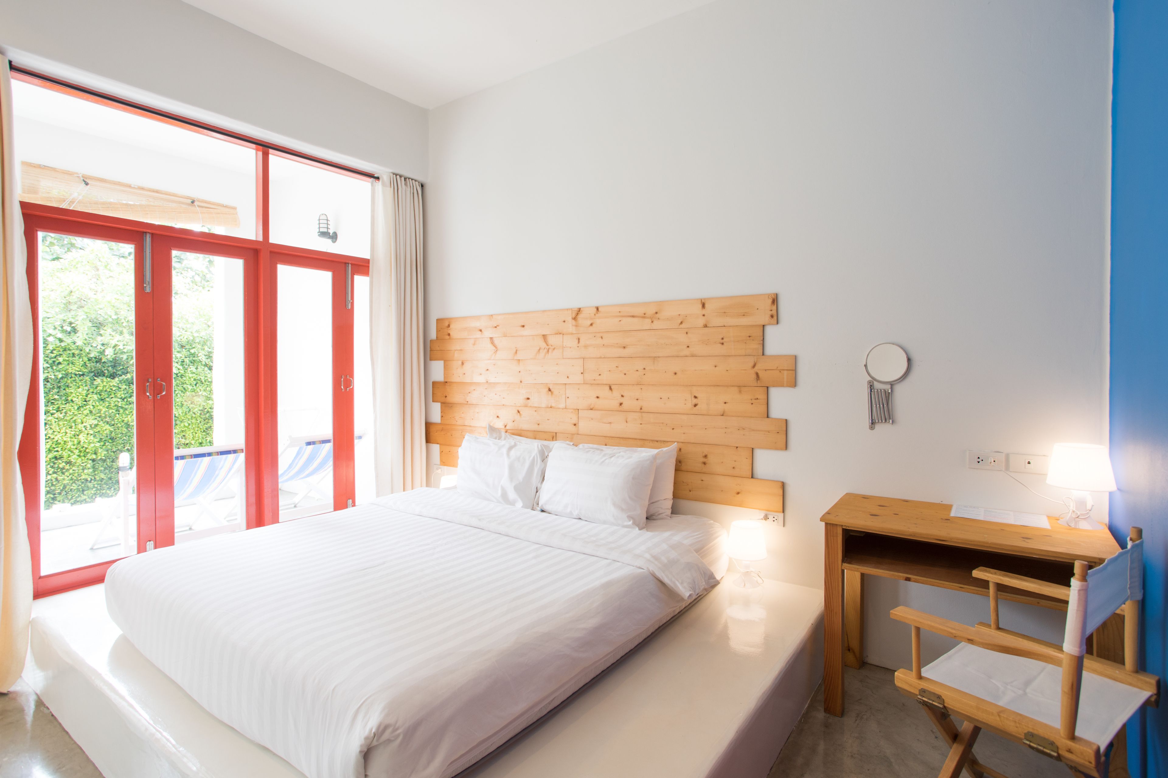 A bedroom with a wooden headboard, a bed with white linens, a small wooden desk with a lamp, and a view through a large window.