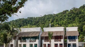 Una hilera de edificios blancos con techos de paja y balcones, frente a una exuberante ladera verde.