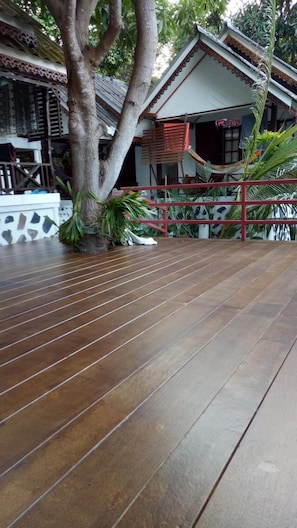 Eine Holzterrasse mit Fliesenboden, umgeben von tropischen Pflanzen und einem Gebäude im traditionellen Stil mit rotem Dach.