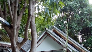 Une terrasse en bois avec un arbre au premier plan, une maison avec une balustrade rouge et une végétation luxuriante en arrière-plan.