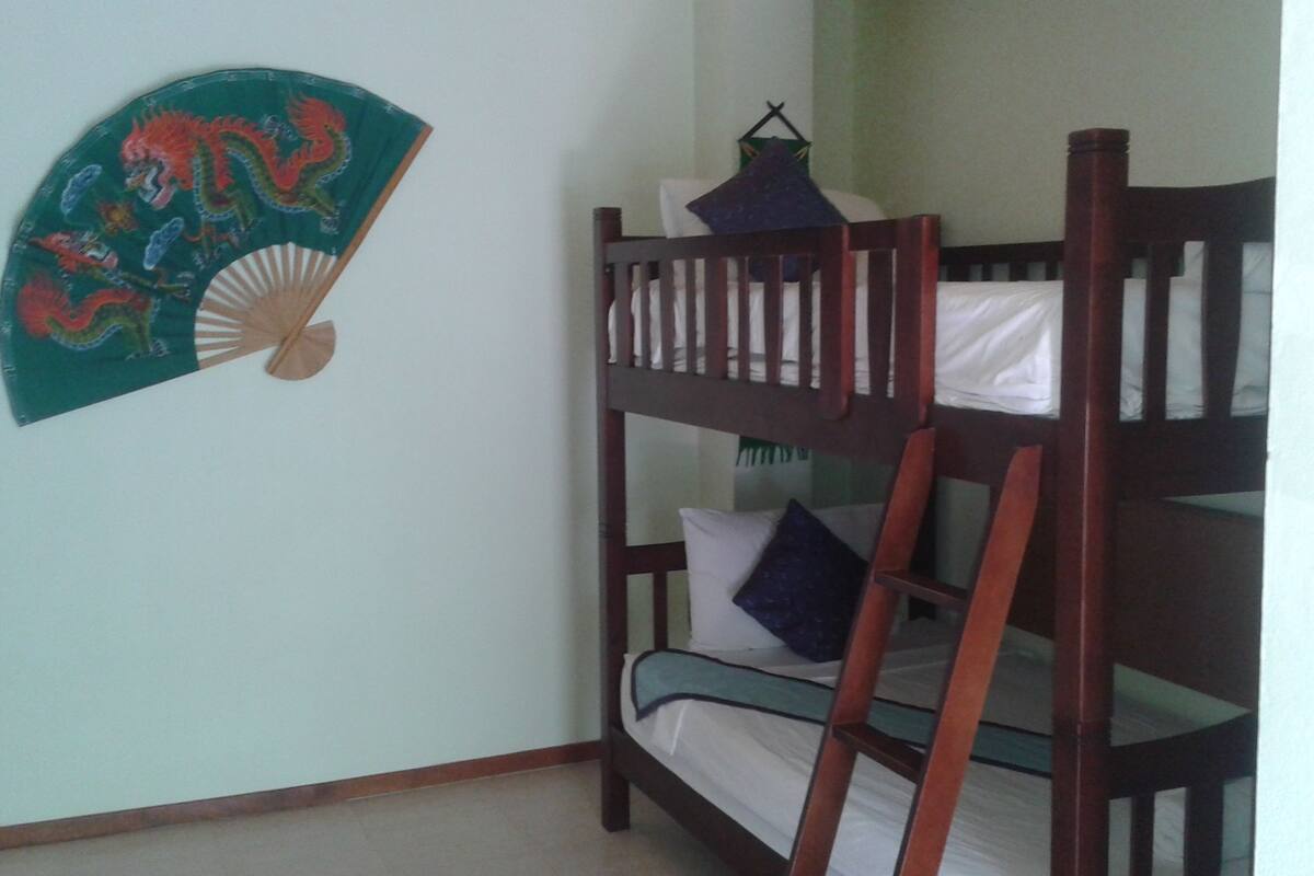 Een stapelbed met houten frame en witte beddengoed, een decoratieve ventilator met een draak erop en een klein hanglampje.