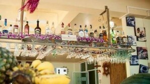 A bar area with bottles and glasses on shelves, a fruit display with bananas and oranges, and a decorative hanging ornament. - VIVA on the Beach Hotel (Ko Pha-ngan)
