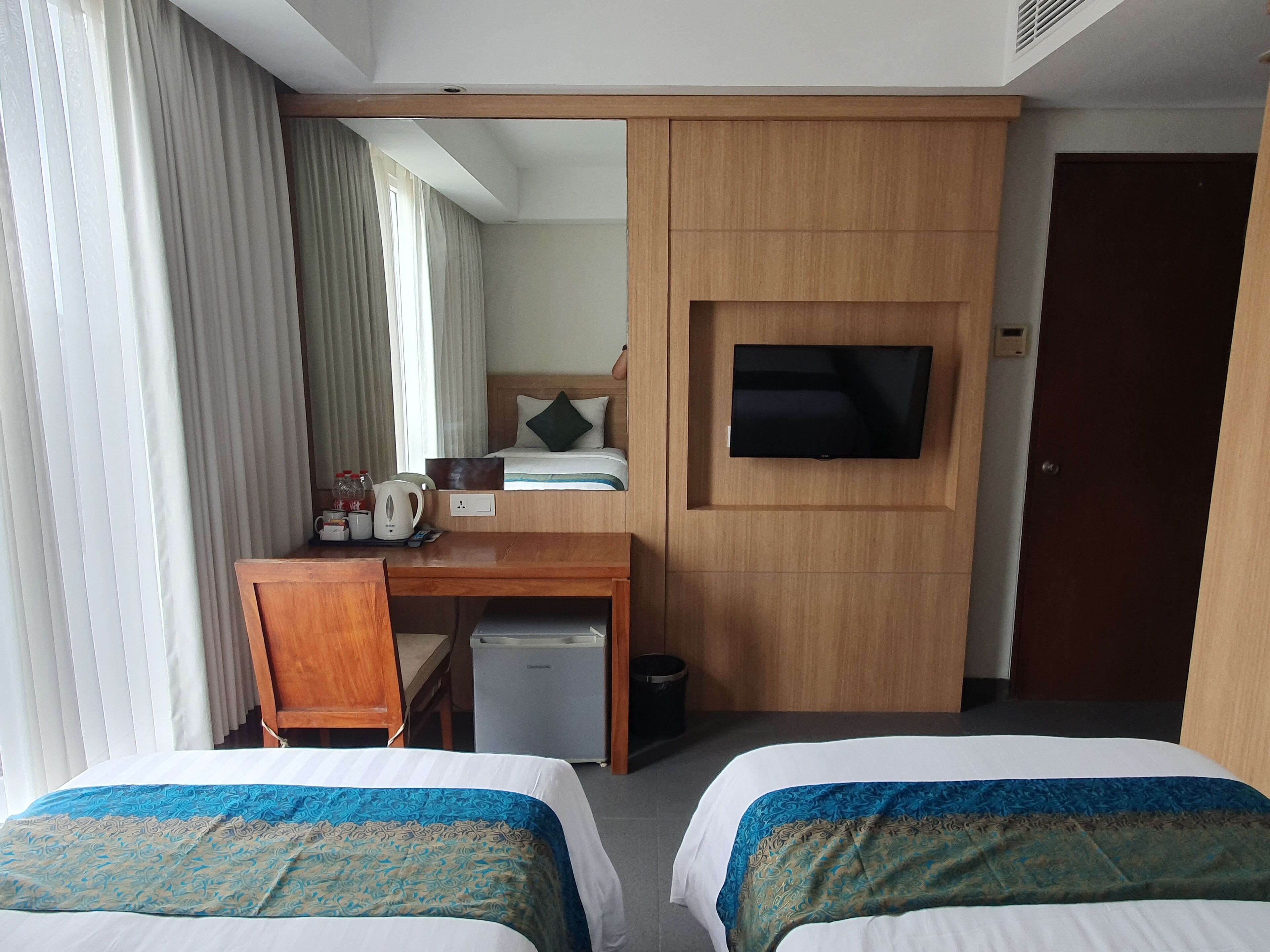 A hotel room with two beds, a desk, a television, and a small kitchenette.