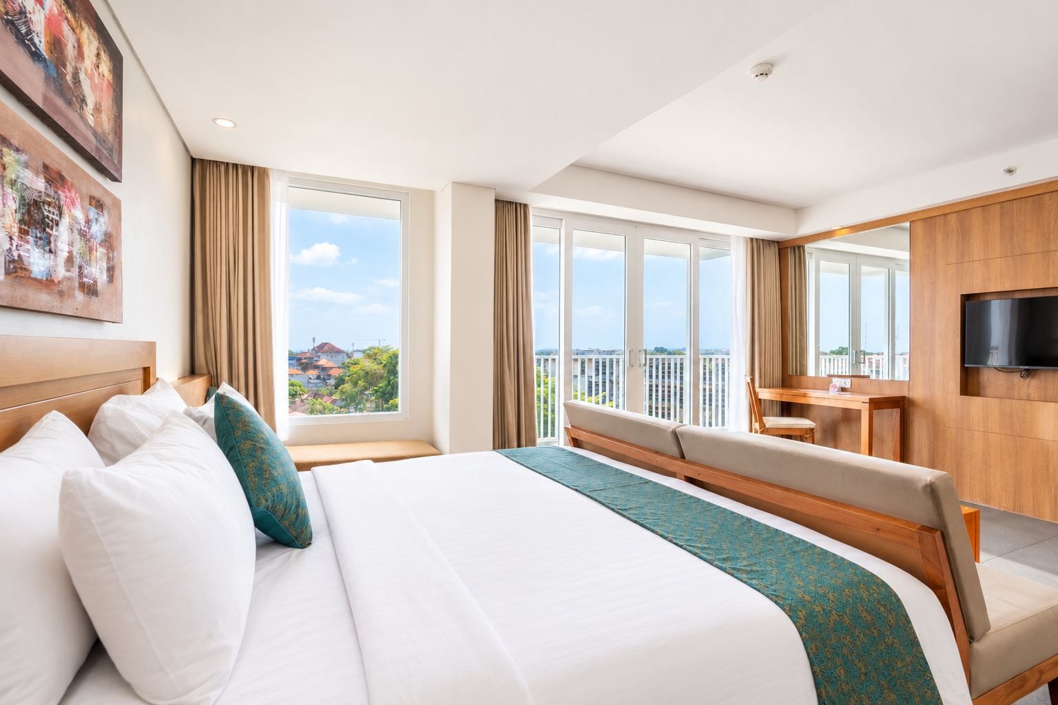 A hotel room with a large bed, a desk, a TV, and a balcony with a city view.