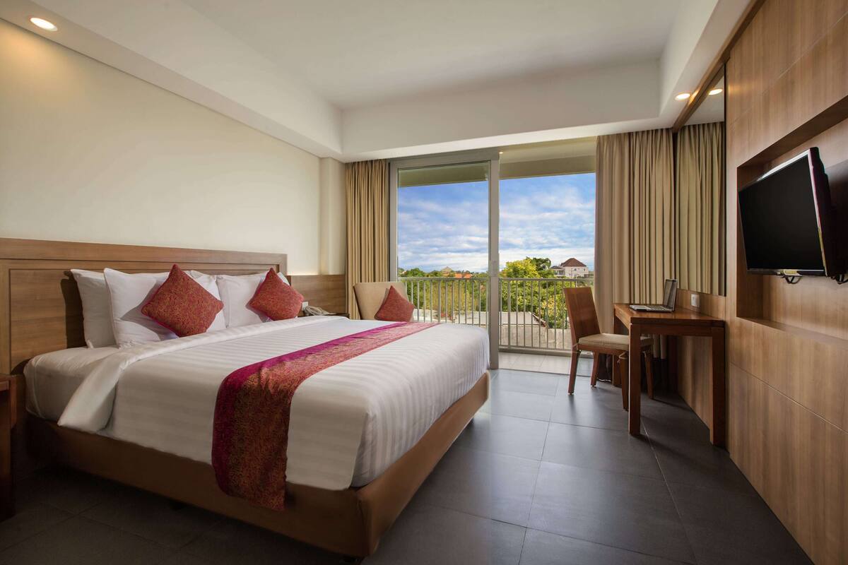 A hotel room with a large bed, a desk, a TV, and a balcony with a view.