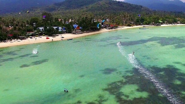 An aerial view of a beach with clear turquoise water, a sandy shore, and a backdrop of mountains. - Dancing Elephant Beach Club - Hostel (Ko Pha-ngan)