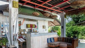 A covered outdoor seating area with a bar, cushioned seating, and hanging baskets. - Dancing Elephant Beach Club - Hostel (Ko Pha-ngan)