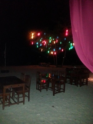 Ein Essbereich im Freien mit Holztischen und -stühlen, umgeben von einem Sandstrand bei Nacht.