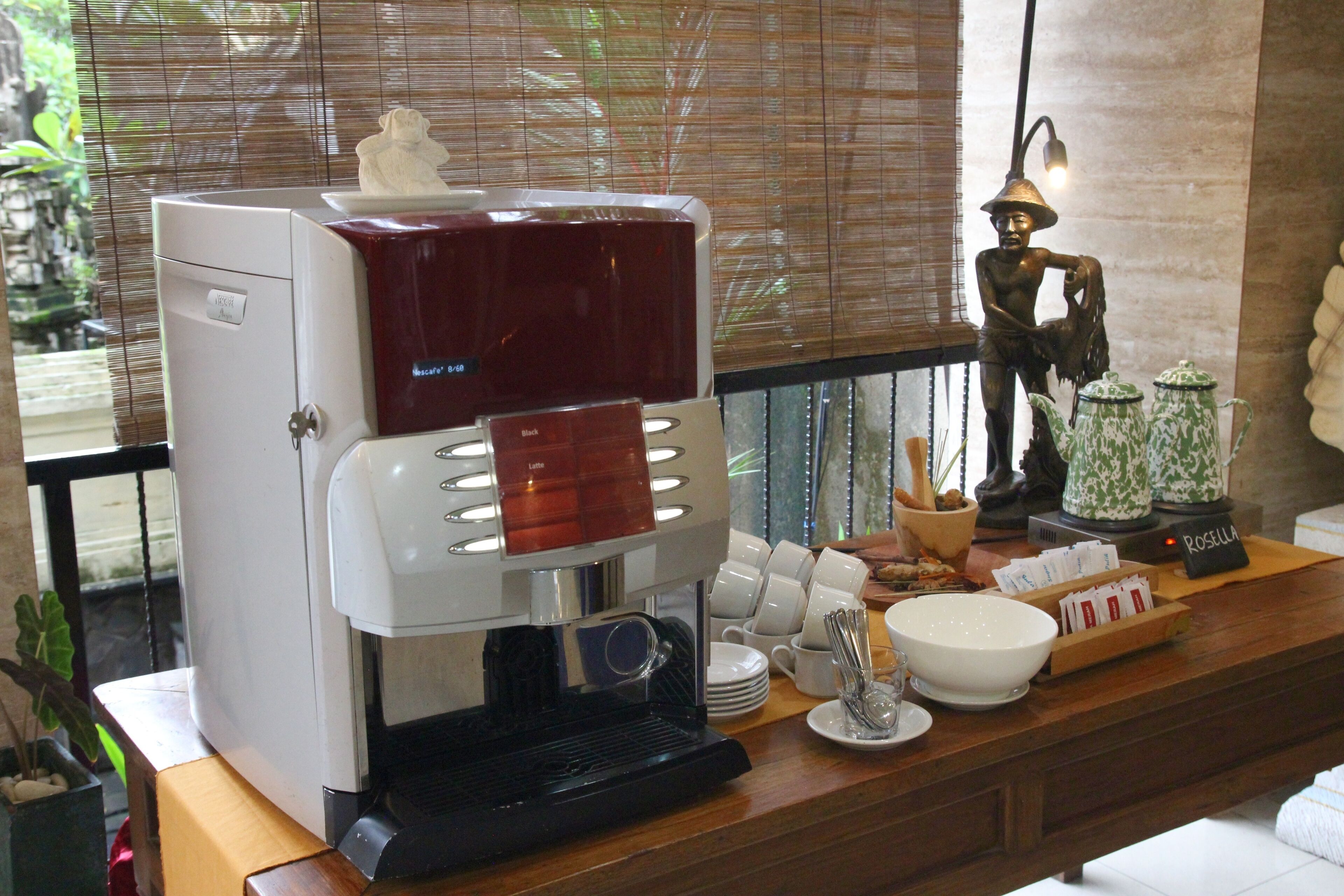 Una cafetera con espumador de leche, una estatua de cerámica y un juego de tazas y platillos blancos sobre una encimera de madera.