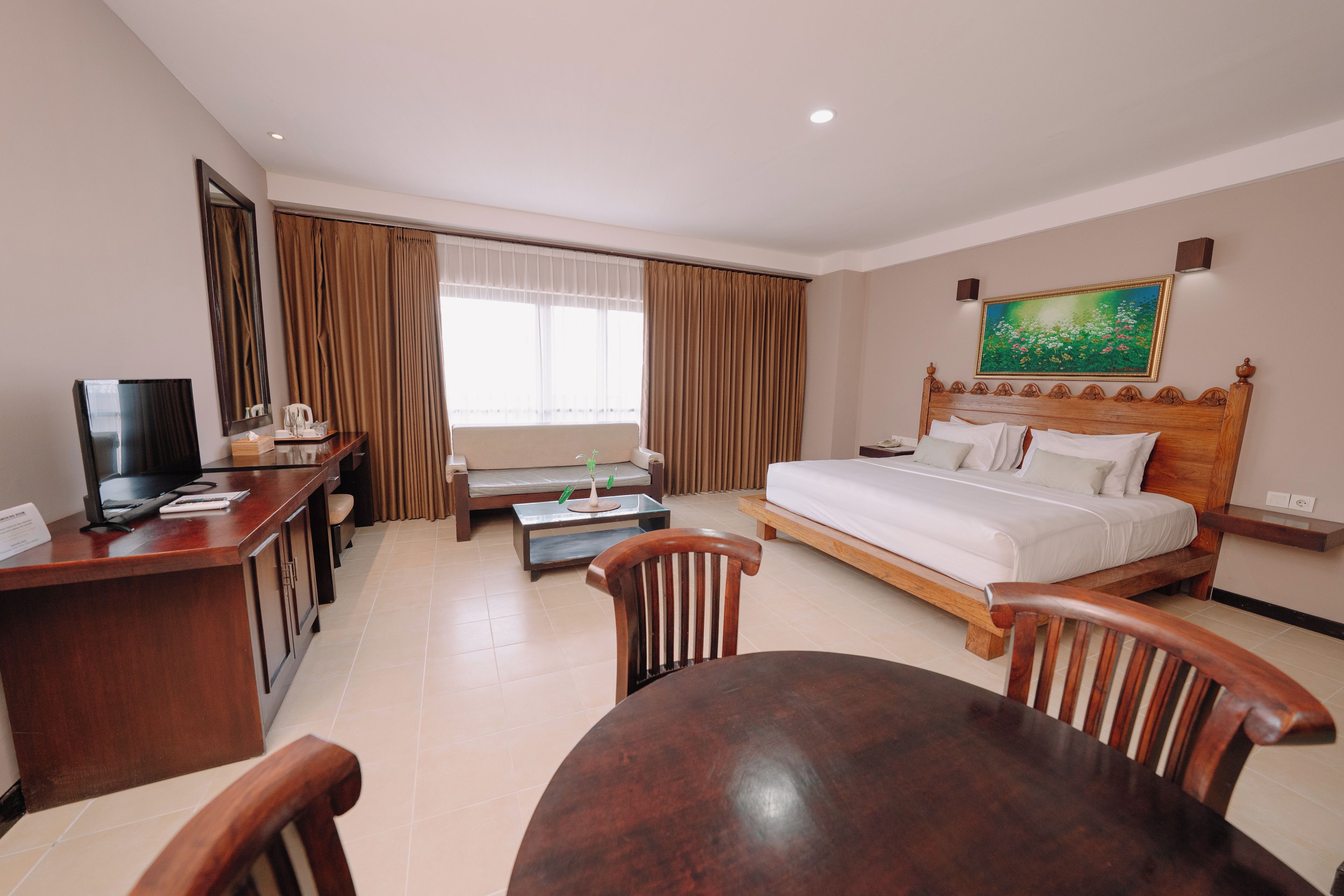 Quarto de hotel com cama, escrivaninha com televisão, mesa de jantar com cadeiras, sofá e janela com cortinas.