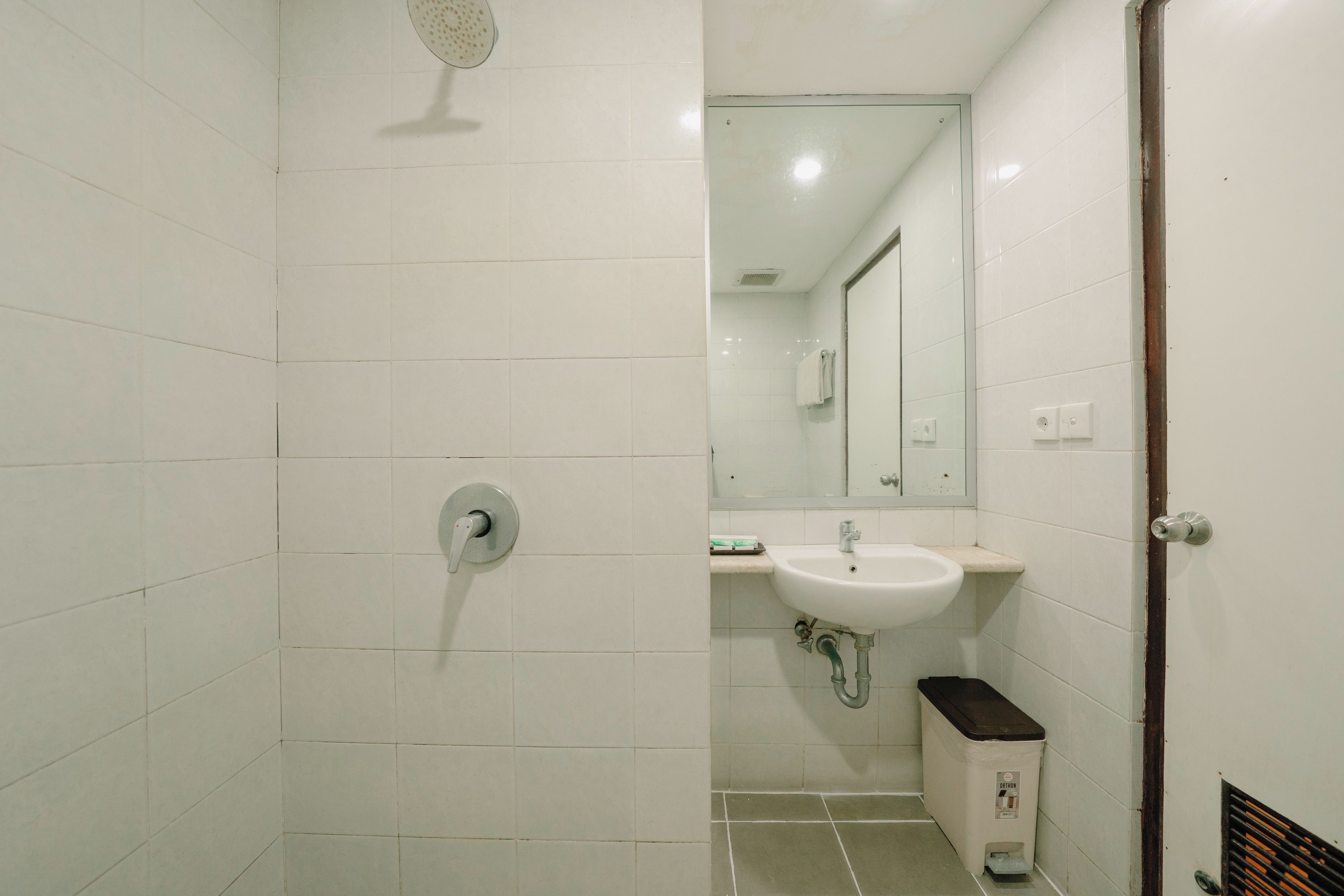 Un baño con azulejos blancos, ducha, lavamanos y cubo de basura.