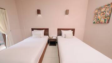 A hotel room with two single beds, a nightstand with a phone and papers, a window with curtains, and a colorful abstract painting on the wall.