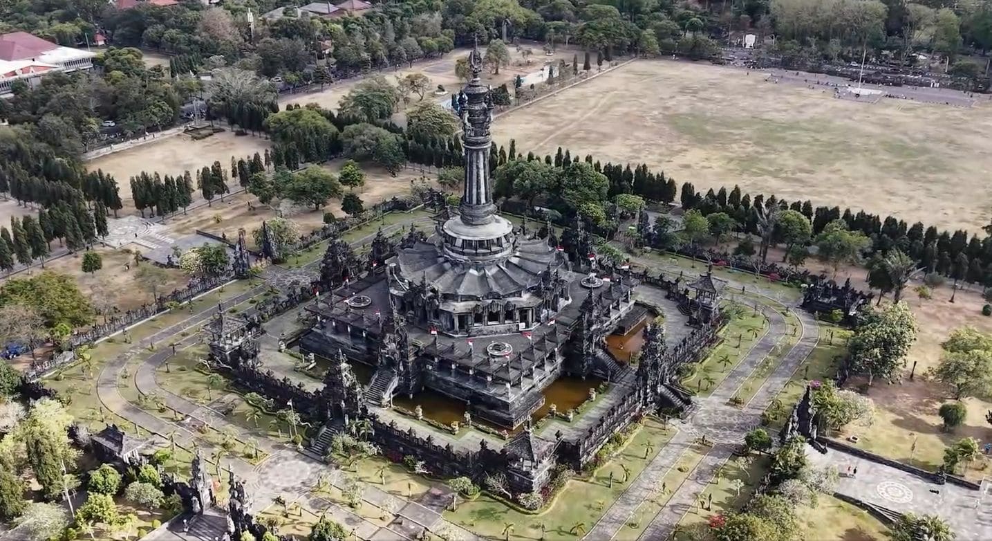 An aerial view of a large, historic temple complex with multiple structures, including a central tower, surrounded by pathways and greenery.
