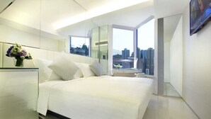 A modern hotel room with a large bed, a glass desk, a vase of flowers, and a city view through a large window.
