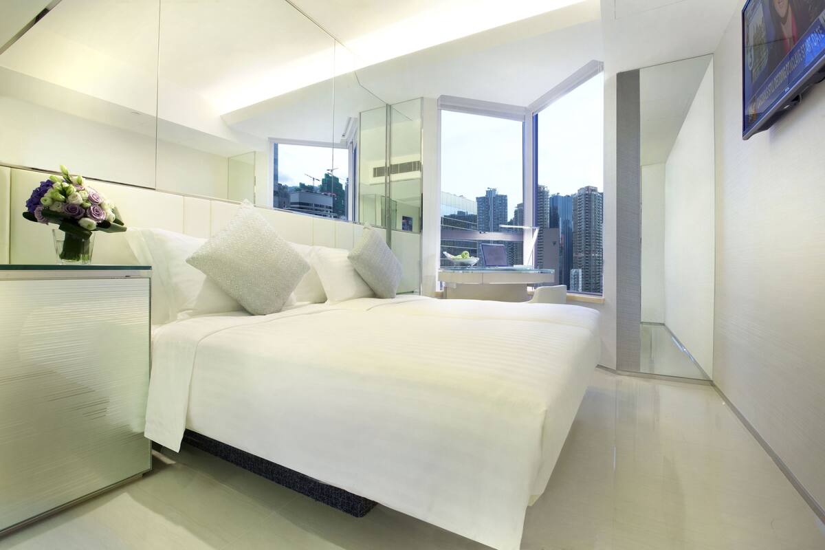 Habitación de hotel moderna con una cama grande, un escritorio de vidrio, un jarrón de flores y vistas a la ciudad a través de un ventanal amplio.