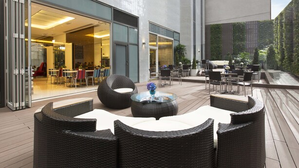 An outdoor seating area with wicker furniture and a glass table, overlooking a modern building with large windows and a patio.
