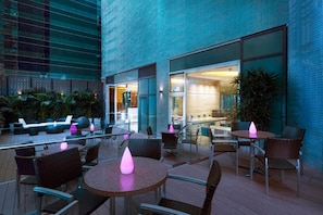 Relax in a comfortable outdoor seating area with round tables and wicker chairs, surrounded by lush greenery and a view of the interior.