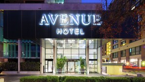 The exterior of the Avenue Hotel with a prominent illuminated sign, a glass entrance, and a well-maintained hedge in front. - Avenue Hotel Canberra (Braddon)