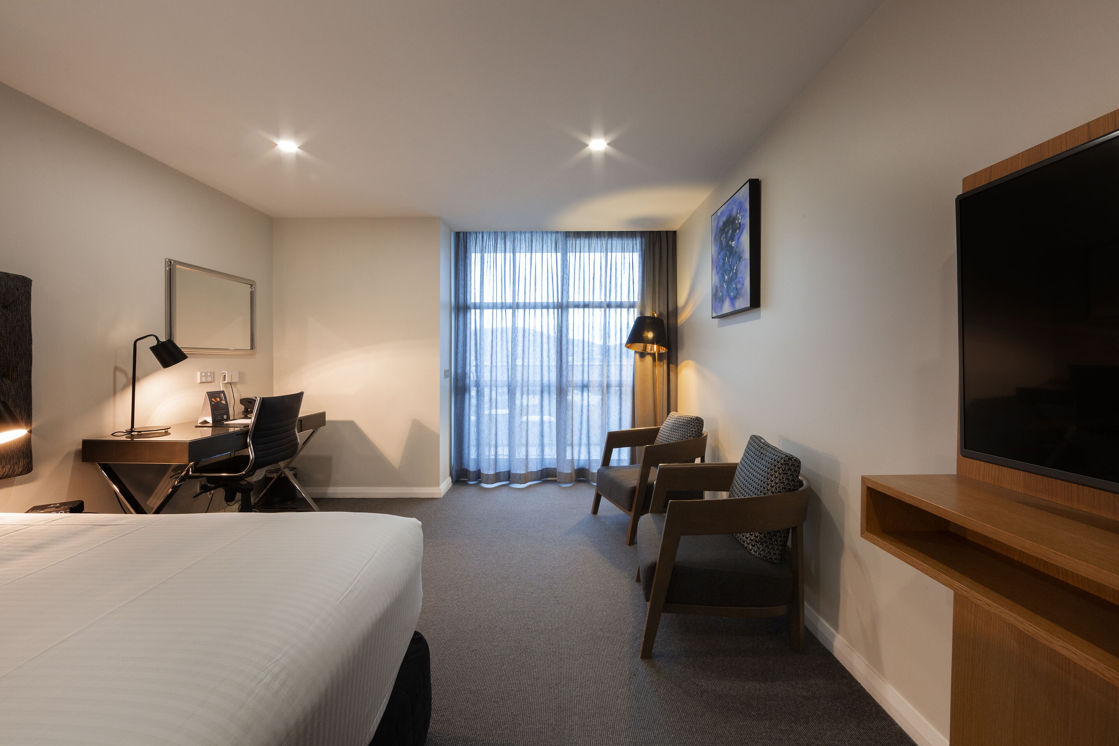 Quarto de hotel com cama, escrivaninha, cadeiras, televisão e uma janela grande com cortinas.