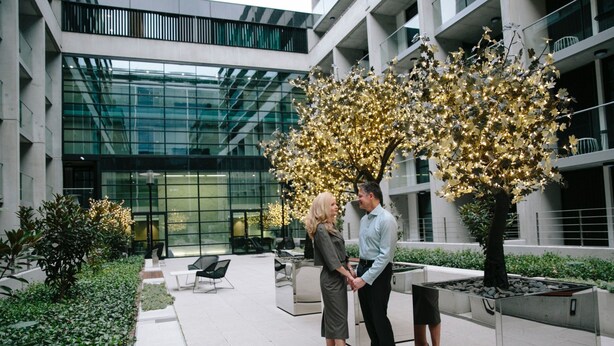A modern indoor space with a glass facade, a central tree with lights, and seating areas. - Avenue Hotel Canberra (Braddon)
