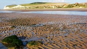 Une plage bordée d’un rivage rocheux, d’où l’on aperçoit, au loin, des maisons situées sur une colline.