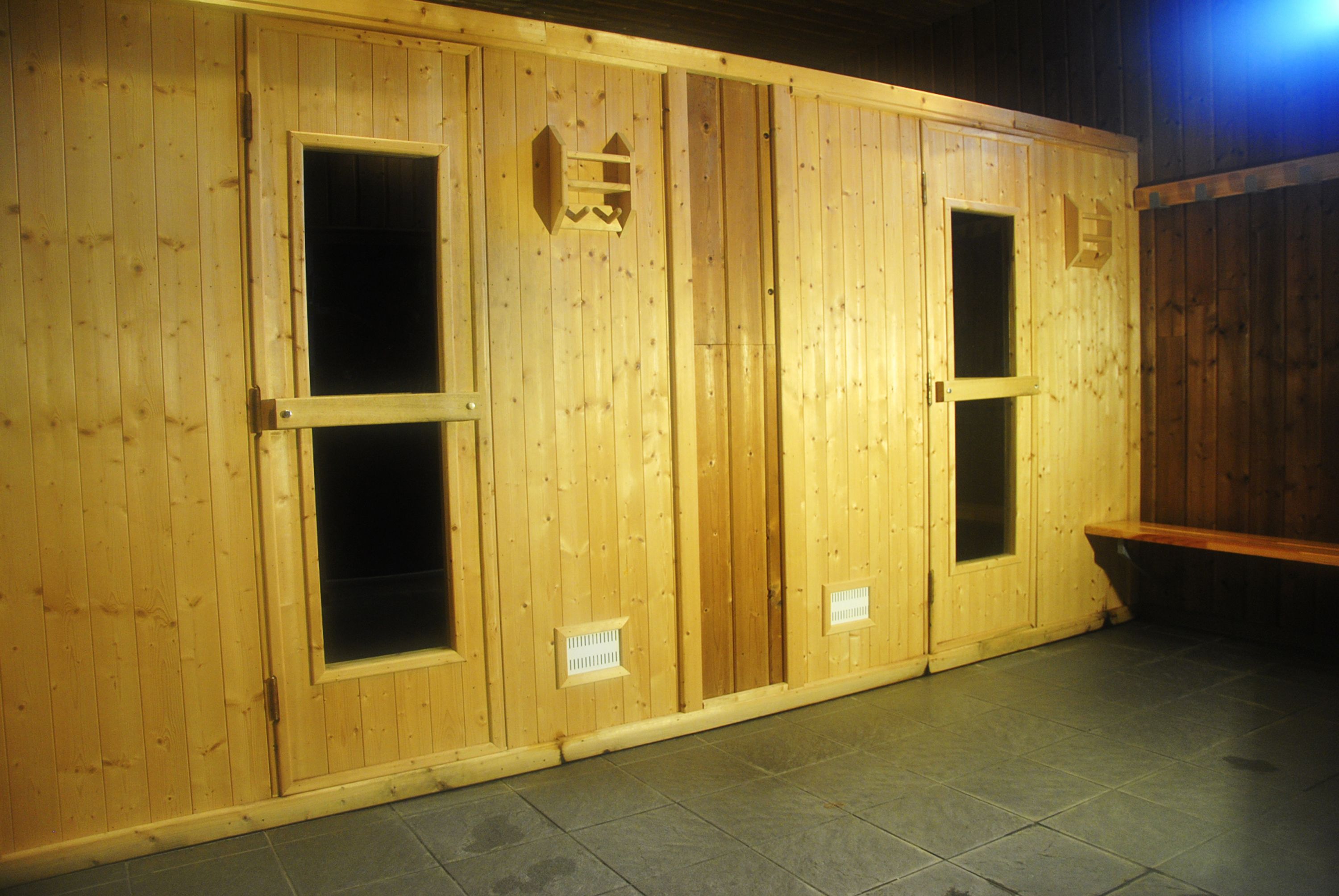 Ein Zimmer mit Holzboden und vier Türen, von denen jede ein kleines Fenster hat. In der Ecke steht eine Bank.