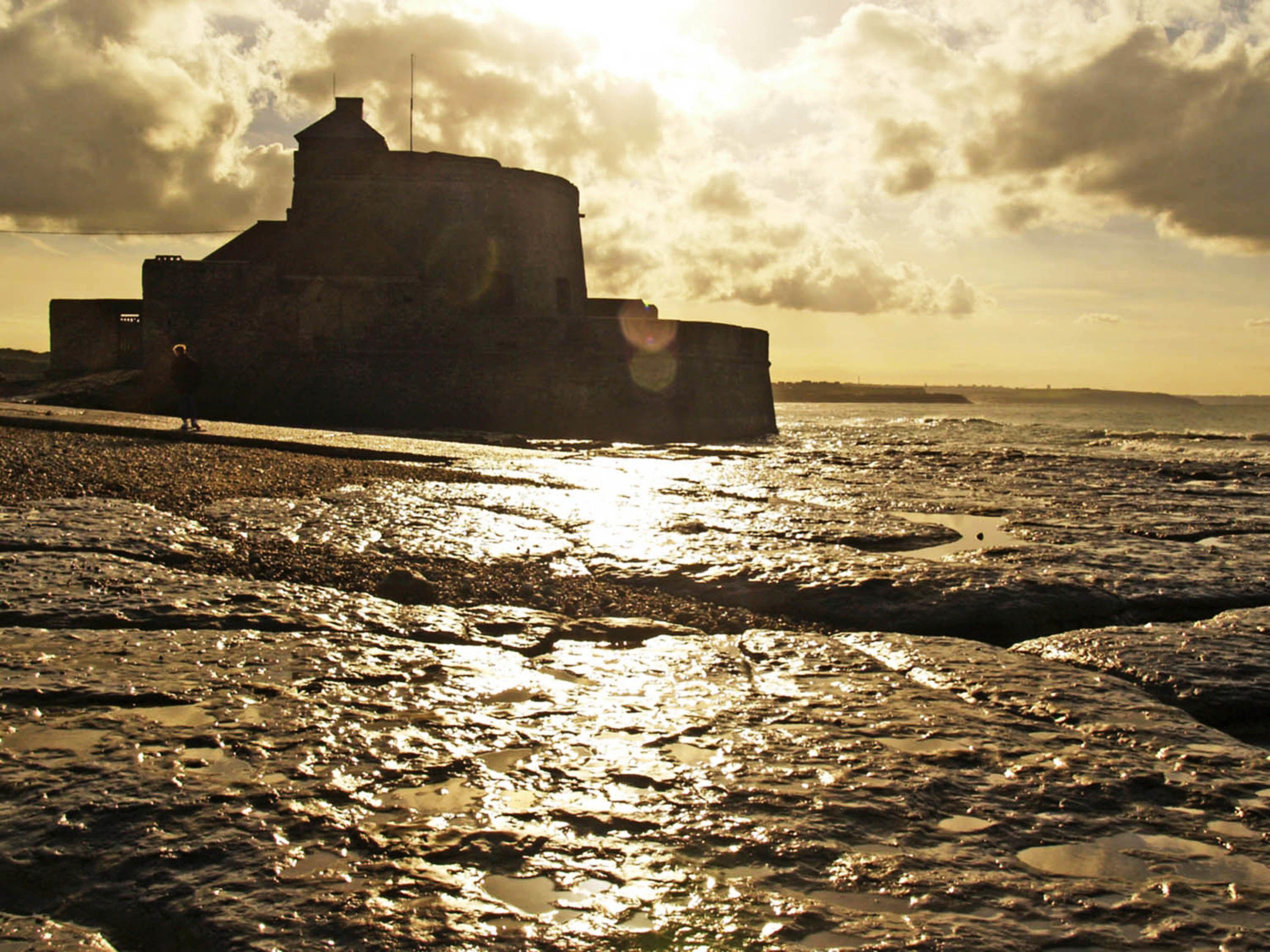 A coastal building with a prominent tower, situated on a rocky shoreline with the sun setting in the background.