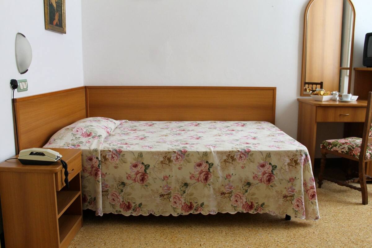 Une chambre avec un lit en bois, une table de chevet avec un téléphone, une chaise, une petite table avec un miroir et une lampe fixée au mur.