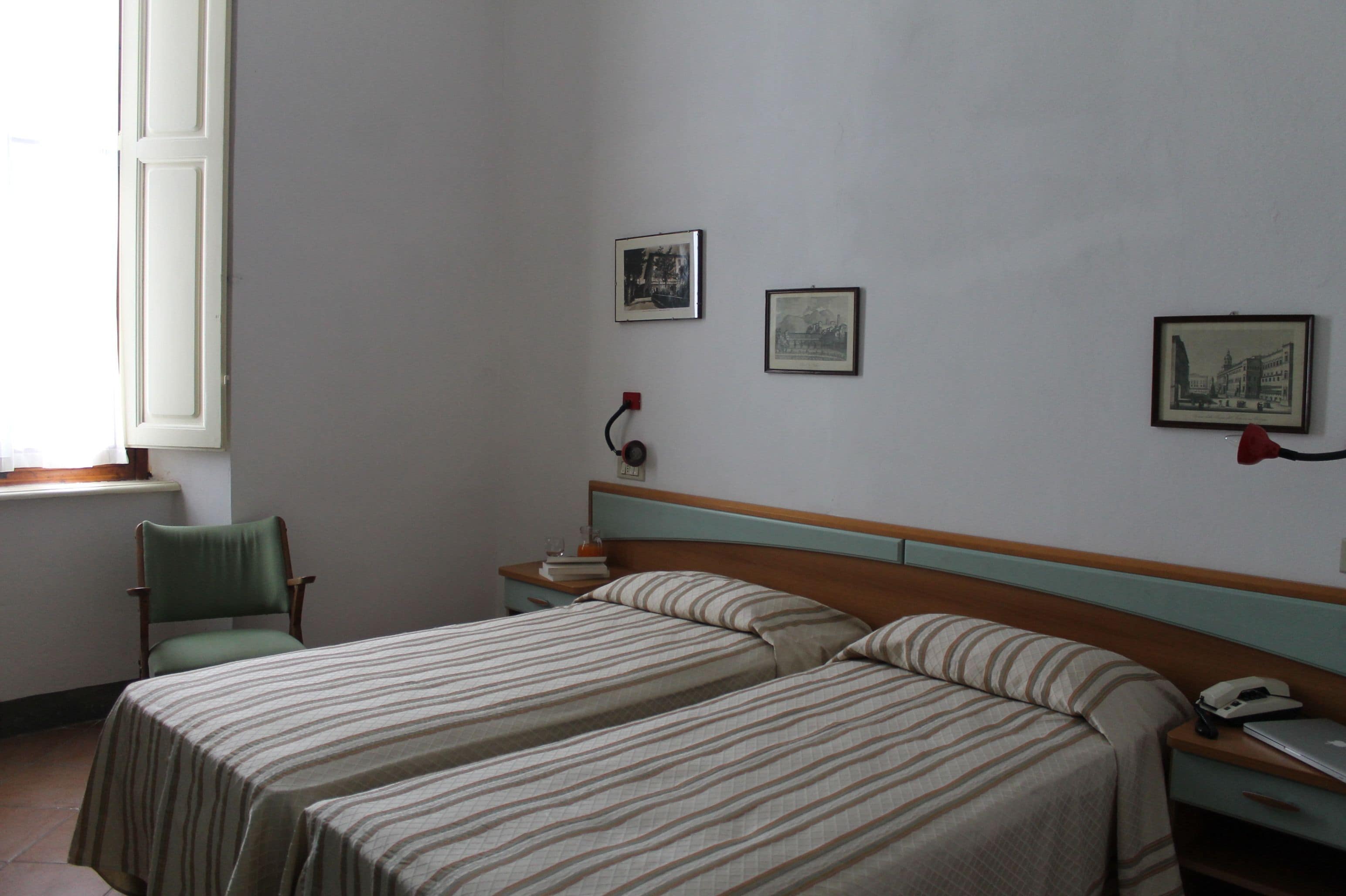 Ein Zimmer mit zwei Betten, einem Sessel, einem Nachttisch mit Lampe und gerahmten Bildern an der Wand.