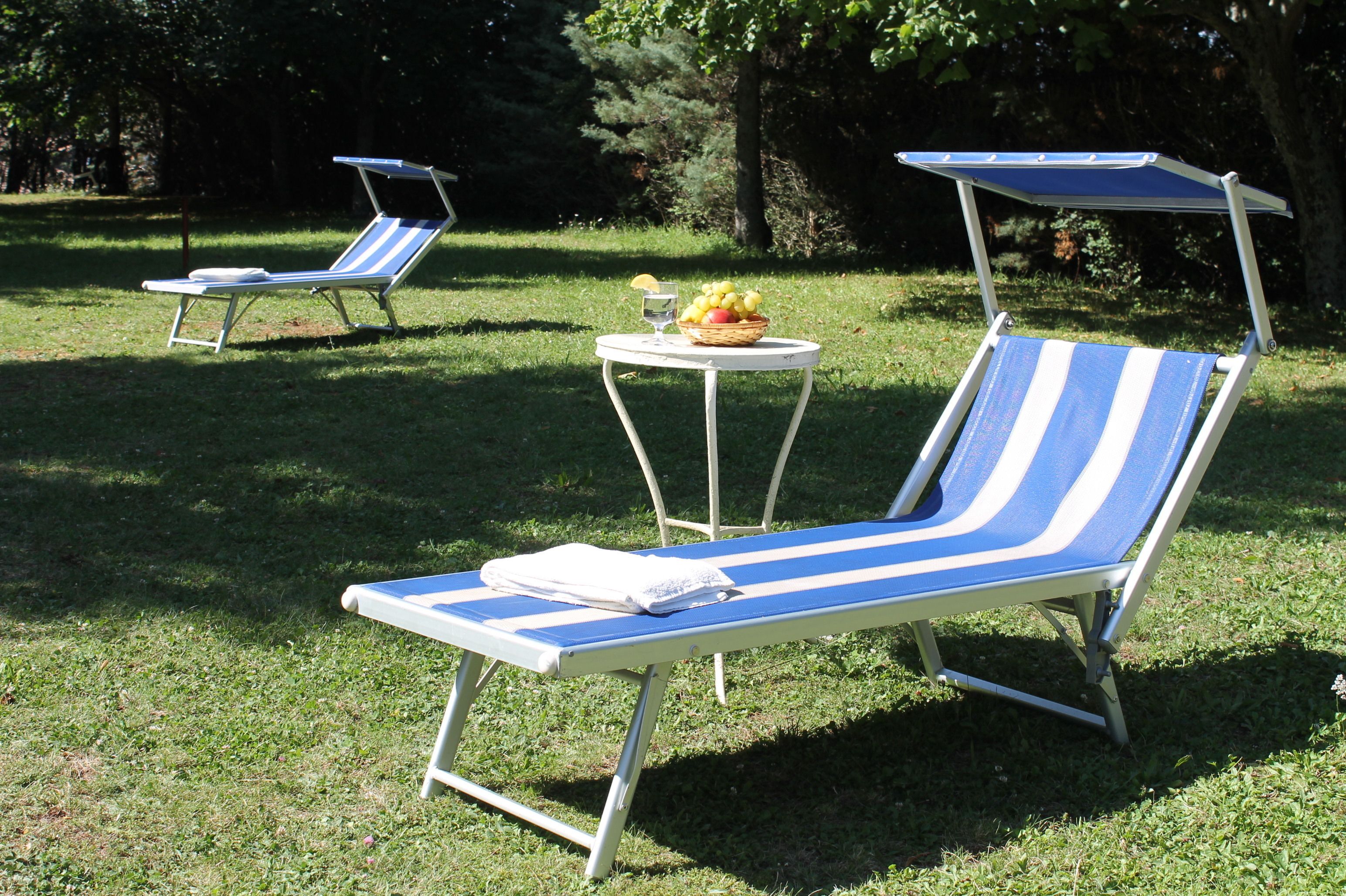 Two blue and white striped lounge chairs with attached umbrellas, a small table with a basket of fruit, and a folded towel on one of the chairs.