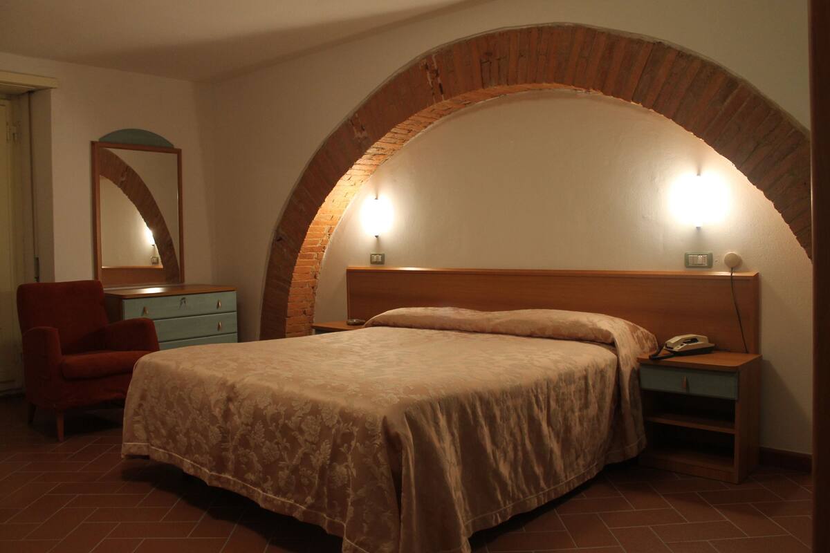 Une chambre avec un grand plafond voûté en briques, un lit recouvert d’une couverture beige, une table de chevet avec une lampe, une chaise et une commode.