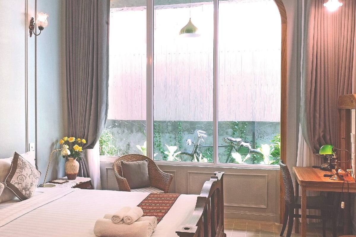 A bedroom with a large window, a bed, a chair, a desk, and a lamp.