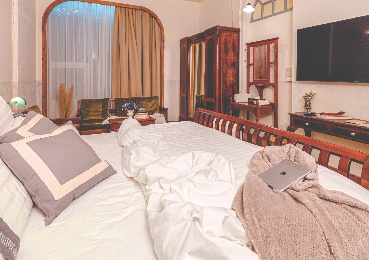 Un dormitorio con cama, computadora portátil, armario de madera, escritorio y una ventana grande con cortinas.