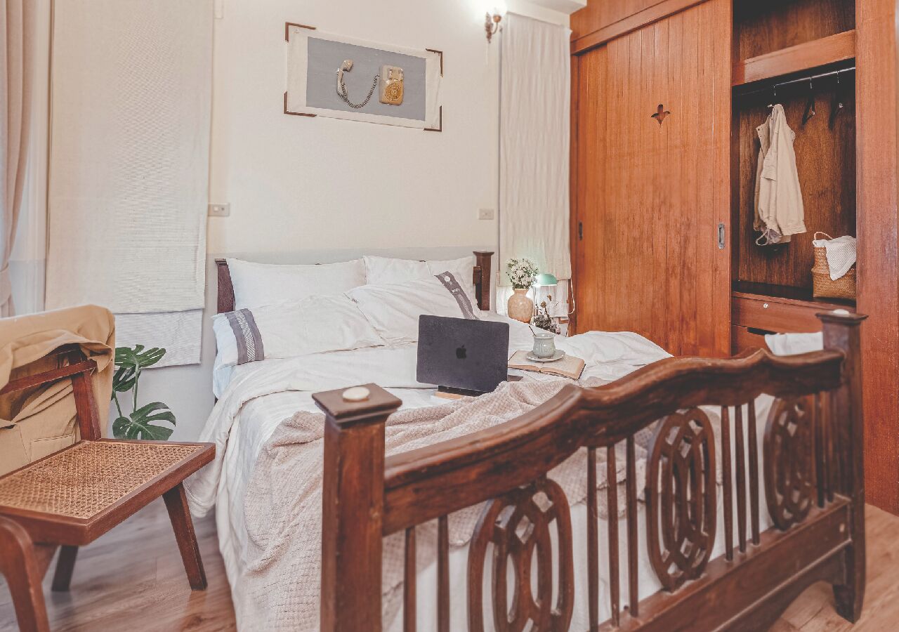 Quarto com cama de madeira, um laptop na cama, guarda-roupa de madeira e uma cadeira de vime.
