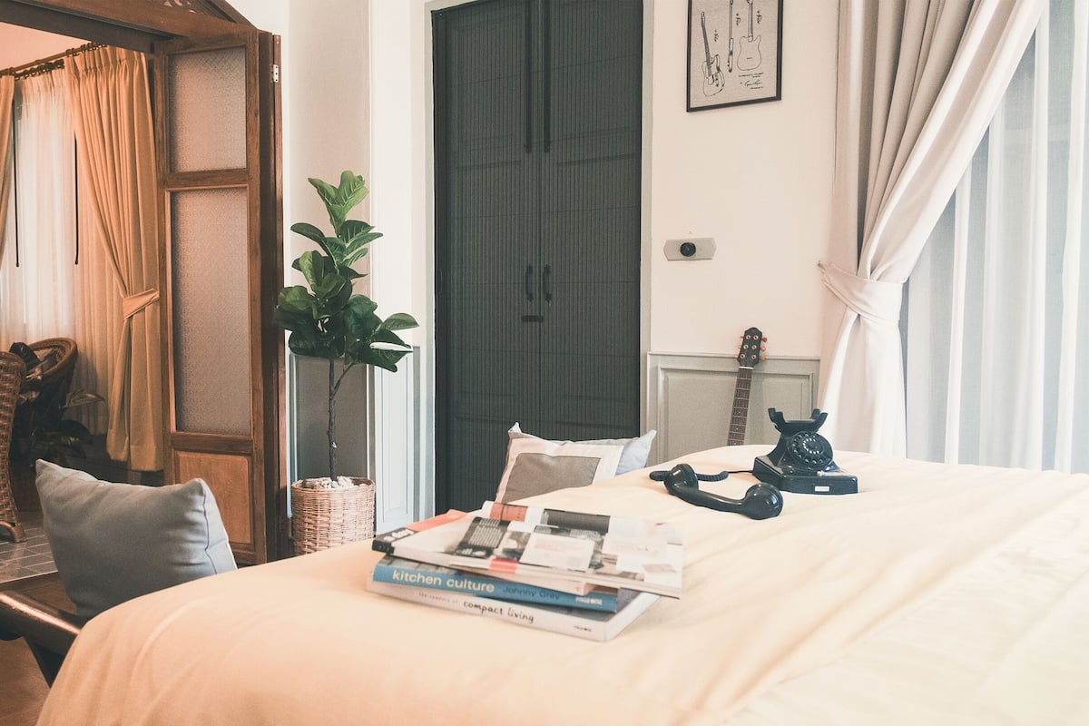 A bedroom with a bed, a bedside table with books, a telephone, a guitar, and a plant.